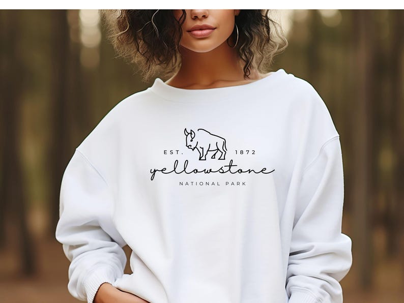 May include: White sweatshirt featuring a black bison graphic and the text "Yellowstone National Park" and "Est. 1872". The top has a crew neck and long sleeves. The design is centrally placed on the chest.