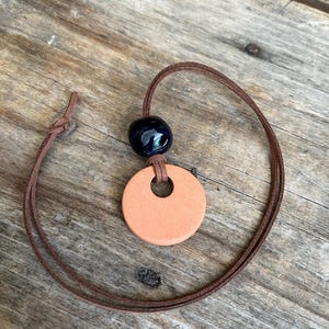 May include: A necklace featuring a round, peach-colored pendant with a central hole, paired with a dark blue bead. The pendant hangs from a brown cord, knotted at the top. The necklace rests on a weathered wooden surface.