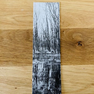 May include: Black and white bookmark featuring a serene forest scene reflected in water. The image shows bare trees and their mirrored reflections, creating a tranquil, nature-themed design. The bookmark is rectangular and rests on a wooden surface.