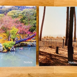Könnte beinhalten: Zwei Notizbücher mit Landschaftsfotografien. Eines zeigt einen japanischen Garten mit Teich, Bäumen und farbenfrohem Laub. Das andere zeigt eine Strandszene mit Bäumen und einem Holzzaun. Beide Notizbücher tragen das Wort "Notes".