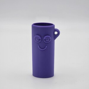 Puede incluir: Un contenedor de silicona morado con asa y una cara sonriente. El contenedor tiene dos ojos y una boca.