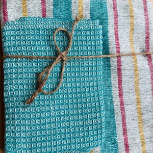 Peut inclure: Une pile de carrés de tissu gaufré bleu sarcelle pliés, attachés avec une fine ficelle brune, posée sur un tissu rectangulaire plus grand présentant des rayures verticales dans des tons bleu sarcelle, jaune, magenta et blanc, à l'aspect texturé.