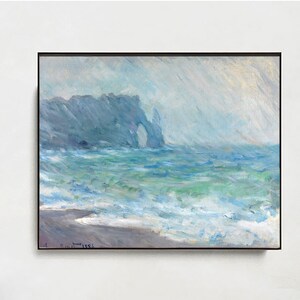 Puede incluir: Una pintura al óleo en un marco negro representa una escena costera con una formación rocosa y olas del océano. La obra de arte se exhibe sobre un banco de madera con patas de metal negro. Un sombrero fedora gris descansa sobre el banco.