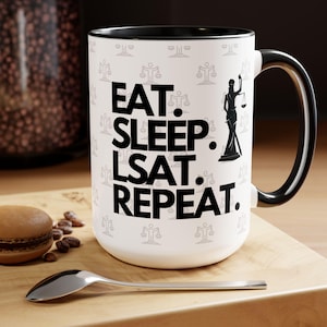 Könnte beinhalten: Eine weiße Keramiktasse mit schwarzem Rand und Henkel. Die Tasse hat ein Muster aus Justizwagen und den Text "EAT. SLEEP. LSAT. REPEAT." in Schwarz.