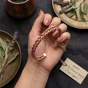 Op de afbeelding: Een koperen gevlochten manchetarmband. De armband wordt in een hand gehouden en heeft een geweven ontwerp. Een label aan de armband luidt "AURA - The Braid of Serenity | Pure Copper."