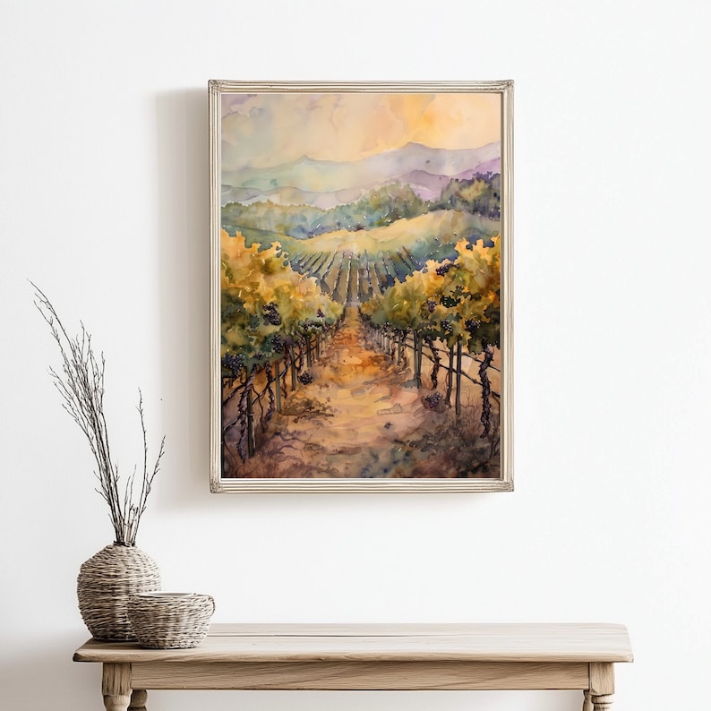 May include: Watercolor painting of a vineyard scene. The artwork depicts rows of grapevines with purple grapes, leading towards distant hills under a warm, golden sky. The painting is framed in a silver-colored frame.