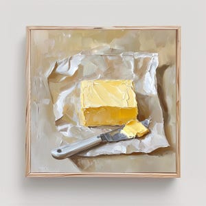 May include: An oil painting of a square block of yellow butter resting on a piece of crumpled white parchment paper. A silver butter knife with a small amount of butter on its blade lies across the paper. The artwork is in a light wood frame.