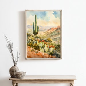 May include: Watercolor painting of a desert landscape featuring a tall saguaro cactus and prickly pear cacti with red flowers. The artwork is framed in a silver-colored frame and depicts a scenic view of the desert with mountains in the background.