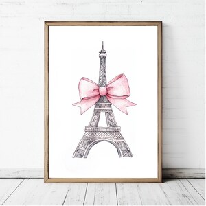 May include: A framed wall art print featuring a black and white ink-style illustration of the Eiffel Tower. A large, soft pink watercolor-style bow is tied around the middle section of the tower structure, centered against a plain white background.