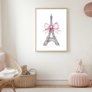 May include: A framed wall art print featuring a detailed black and white sketch of the Eiffel Tower. The tower is adorned with a large, soft pink watercolor-style bow tied around its midsection, centered against a plain white background.