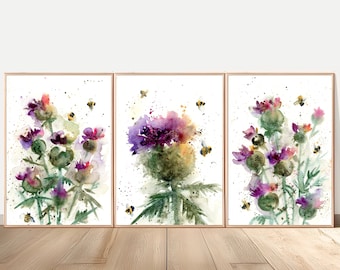 May include: A triptych of three framed watercolor paintings featuring purple and green thistle flowers with scattered paint splatters and small yellow and black bees. Each rectangular print is encased in a thin light wood frame.