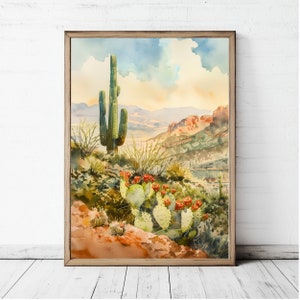 May include: Watercolor painting of a desert landscape featuring a tall saguaro cactus and prickly pear cacti with red flowers. The artwork is framed in a wooden frame and depicts a scenic view of the desert with mountains and a cloudy sky.