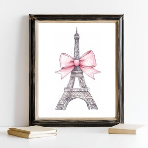May include: A framed wall art print featuring a black and white ink illustration of the Eiffel Tower adorned with a large, soft pink watercolor bow tied around its midsection, presented in a distressed black wooden frame with gold inner trim.