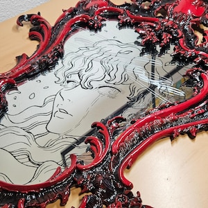 Puede incluir: Espejo ornamentado con marco rojo y negro. El marco presenta un diseño esculpido y detallado. La superficie del espejo refleja una ilustración en blanco y negro de un rostro con cabello suelto.