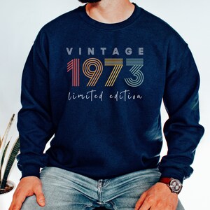 Puede incluir: Una sudadera azul marino con el texto "Vintage 1973 edici&oacute;n limitada" en un esquema de colores arco&iacute;ris.