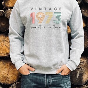 Puede incluir: Una sudadera gris con el texto "Vintage 1973 edici&oacute;n limitada" en colores arco&iacute;ris. Los n&uacute;meros est&aacute;n estilizados con rayas.