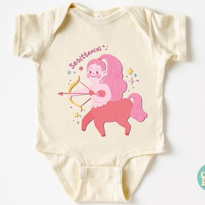 May include: A cream-coloured baby onesie featuring a pink and red illustration of a Sagittarius zodiac sign. The illustration depicts a centaur with a bow and arrow, surrounded by stars.