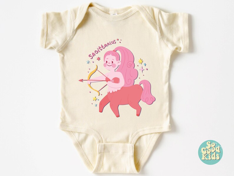 May include: A cream-coloured baby onesie featuring a pink and red illustration of a Sagittarius zodiac sign. The illustration depicts a centaur with a bow and arrow, surrounded by stars.
