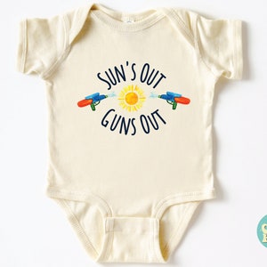 May include: Cream-colored baby onesie with the text "SUN'S OUT GUNS OUT" in navy blue. The design features a yellow sun and two water guns spraying water. A fun summer-themed outfit for babies.