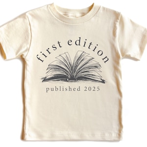May include: A cream-colored short-sleeved t-shirt with a black and white illustration of an open book. The text "first edition published 2025" is printed on the shirt.