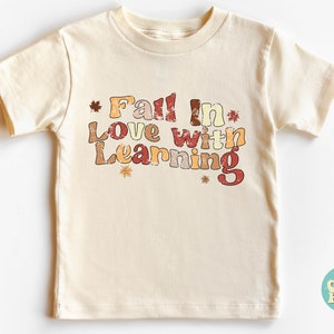 Może przedstawiać: Kremowa koszulka z kr&oacute;tkim rękawem z napisem "Fall in Love with Learning" w stylu vintage z jesiennymi liśćmi. Tekst jest w odcieniach pomarańczowego, czerwonego i brązowego.