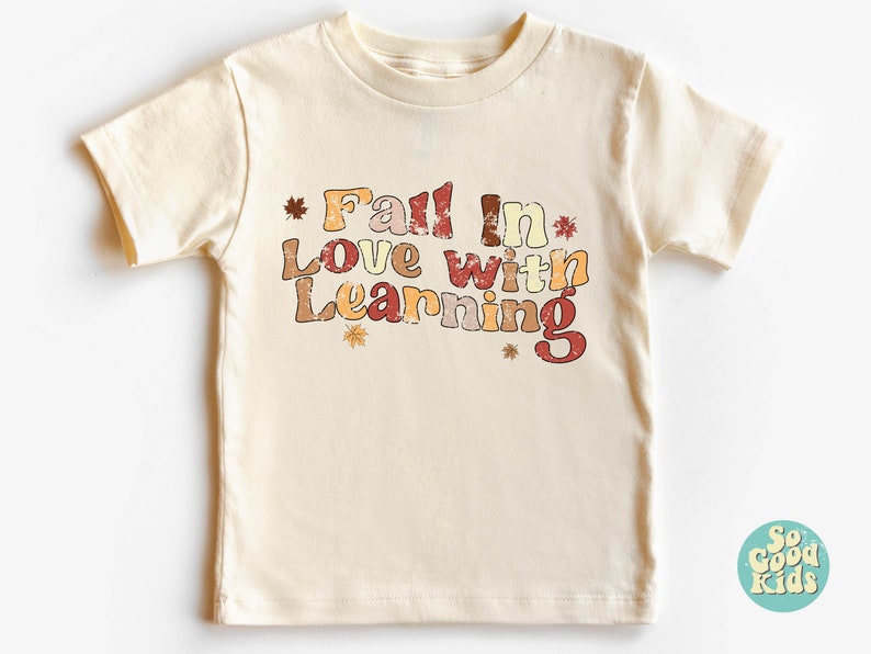 Może przedstawiać: Kremowa koszulka z kr&oacute;tkim rękawem z napisem "Fall in Love with Learning" w stylu vintage z jesiennymi liśćmi. Tekst jest w odcieniach pomarańczowego, czerwonego i brązowego.
