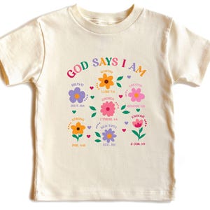May include: Cream-colored t-shirt with the words "GOD SAYS I AM" in a rainbow arc above colorful flower graphics. Each flower has a word like "BRAVE" or "WORTHY". The shirt is from So Good Kids.