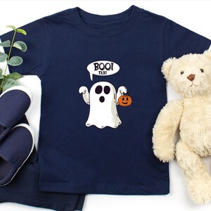Peut inclure: Un t-shirt bleu marine avec un fant&ocirc;me blanc tenant une petite citrouille orange. Le fant&ocirc;me a une bulle de dialogue qui dit "Boo! Yahi!"