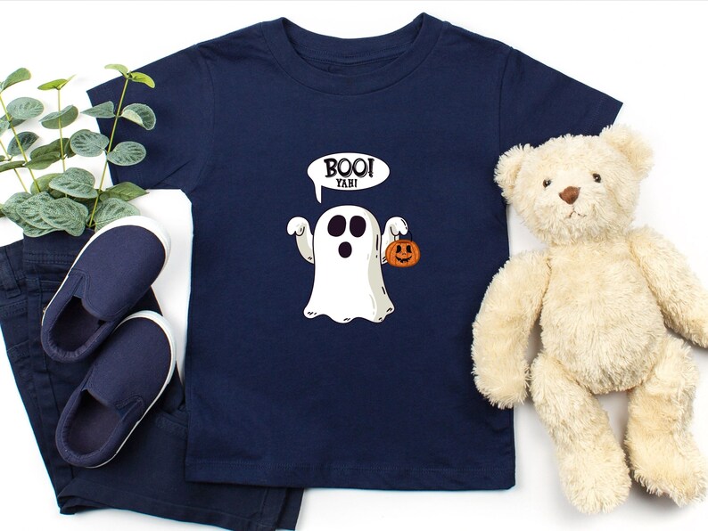 Peut inclure: Un t-shirt bleu marine avec un fant&ocirc;me blanc tenant une petite citrouille orange. Le fant&ocirc;me a une bulle de dialogue qui dit "Boo! Yahi!"