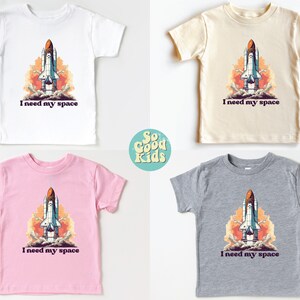 K&ouml;nnte beinhalten: Vier T-Shirts in Kindergr&ouml;&szlig;e in Wei&szlig;, Beige, Rosa und Grau mit einer Grafik eines startenden Raumschiffs. Der Text "Ich brauche meinen Platz" ist unter der Grafik gedruckt.