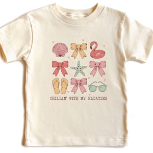 May include: Cream-colored t-shirt featuring a collection of summer-themed illustrations. The design includes a seashell, bows, a flamingo float, a starfish, flip-flops, sunglasses, and the phrase "Chillin' with my floaties."
