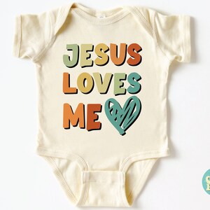 Puede incluir: Un body de beb&eacute; de color crema con el texto "JESUS LOVES ME" en letras retro de colores y un coraz&oacute;n verde.