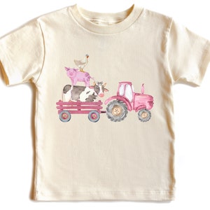 May include: Cream-colored t-shirt featuring a watercolor illustration of a pink tractor pulling a wagon. The wagon carries a cow, a pig, and a chicken. The design has a farm theme.