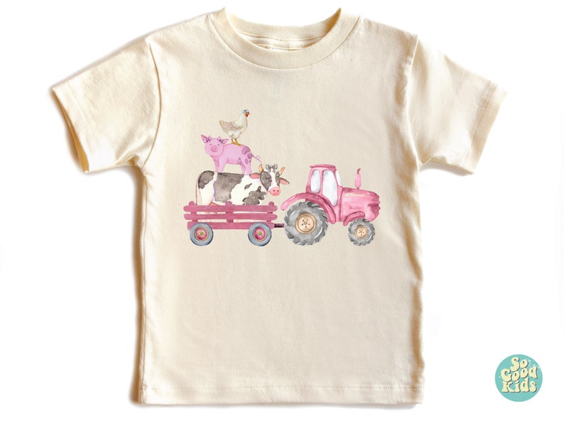 May include: Cream-colored t-shirt featuring a watercolor illustration of a pink tractor pulling a wagon. The wagon carries a cow, a pig, and a chicken. The design has a farm theme.