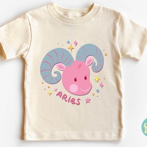 May include: A cream-coloured t-shirt with a pink and blue cartoon ram's head with the word "Aries" printed below it.