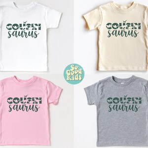 May include: Four toddler-sized t-shirts in different colors with the text "COUSIN saurus" in green. The text is made up of dinosaur silhouettes. The shirts are white, beige, pink, and gray.