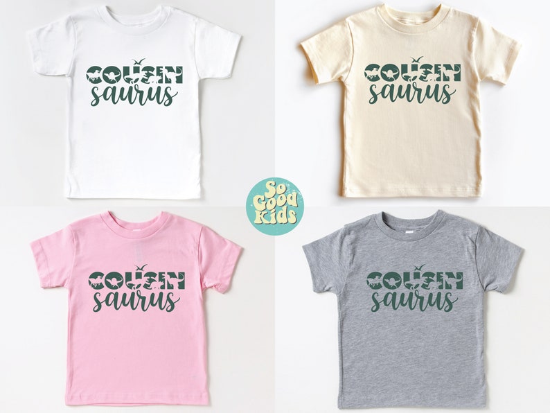 May include: Four toddler-sized t-shirts in different colors with the text "COUSIN saurus" in green. The text is made up of dinosaur silhouettes. The shirts are white, beige, pink, and gray.