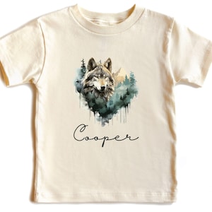 May include: Cream-colored short-sleeved t-shirt featuring a watercolor illustration of a wolf's head surrounded by a forest scene. The name "Cooper" is written in cursive below the image. The shirt has a round neckline.