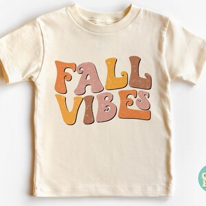 Puede incluir: Una camiseta de manga corta de color crema con el texto "FALL VIBES" en letras de estilo retro de color naranja, marr&oacute;n y rosa.