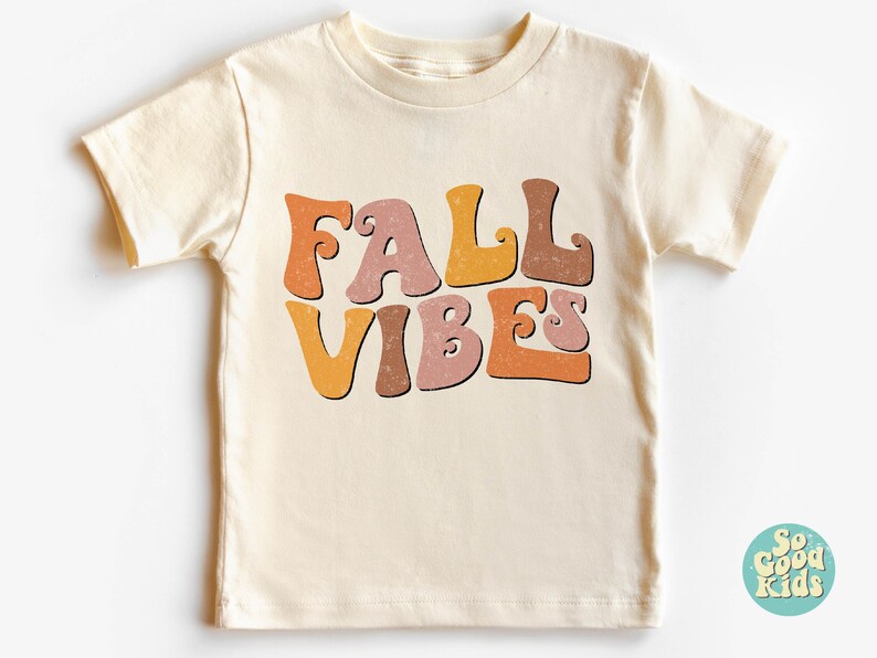 Puede incluir: Una camiseta de manga corta de color crema con el texto "FALL VIBES" en letras de estilo retro de color naranja, marr&oacute;n y rosa.
