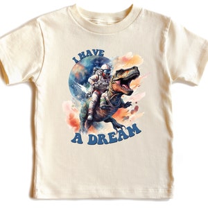 Pode incluir: Uma camiseta bege claro com um gr&aacute;fico de um astronauta montando um dinossauro no espa&ccedil;o. O texto "I HAVE A DREAM" &eacute; impresso em azul abaixo do gr&aacute;fico.