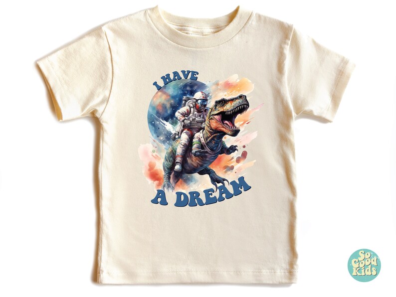 Pode incluir: Uma camiseta bege claro com um gr&aacute;fico de um astronauta montando um dinossauro no espa&ccedil;o. O texto "I HAVE A DREAM" &eacute; impresso em azul abaixo do gr&aacute;fico.