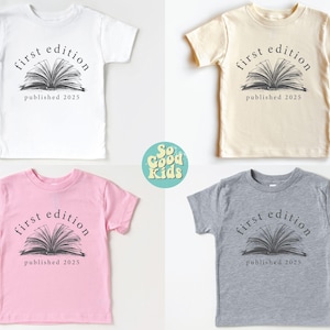May include: Four toddler-sized t-shirts in white, beige, pink, and gray. Each shirt features a black and white graphic of an open book with the text "first edition published 2025".
