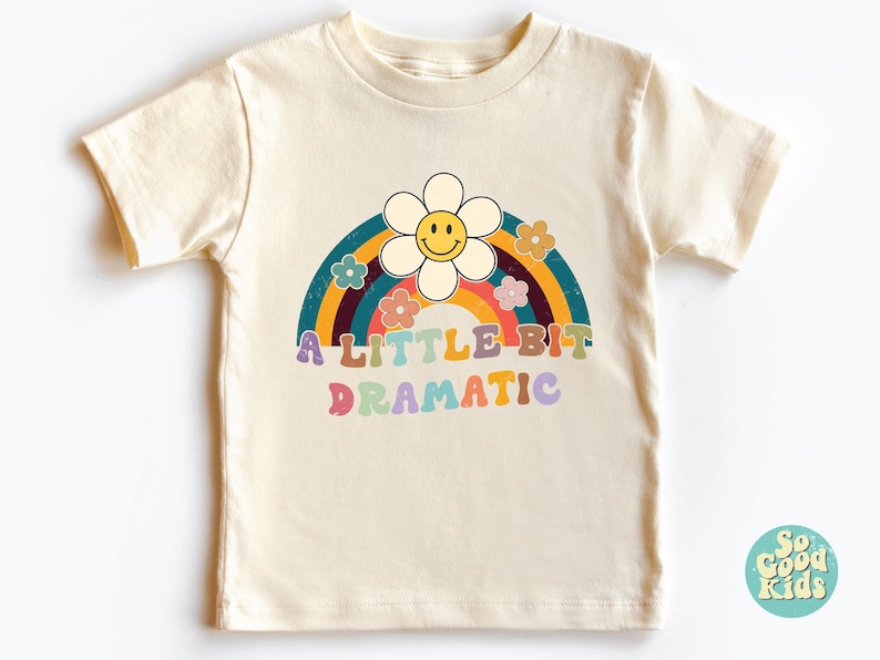 Pode incluir: Uma camiseta bege claro com um arco-&iacute;ris e uma margarida com um rosto sorridente. O texto "A LITTLE BIT DRAMATIC" &eacute; impresso em letras coloridas abaixo do arco-&iacute;ris.