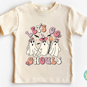 May include: A cream colored t-shirt with a graphic of three ghosts. The ghosts are wearing flowers, a witch hat, and a bow. The text "Let's Go Ghouls" is written in a retro font.