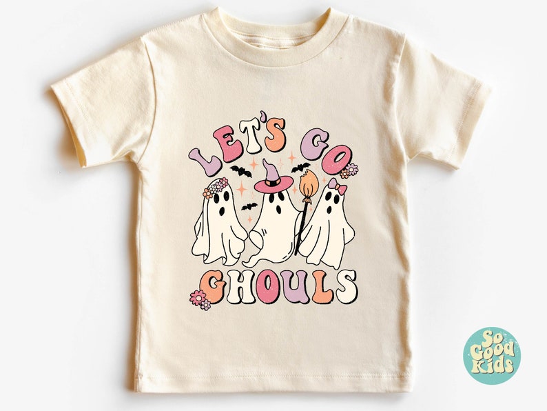 May include: A cream colored t-shirt with a graphic of three ghosts. The ghosts are wearing flowers, a witch hat, and a bow. The text "Let's Go Ghouls" is written in a retro font.