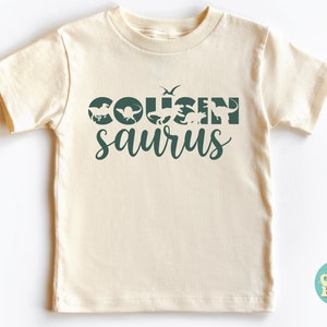 May include: A cream colored t-shirt with a green graphic that says "COUSINsaururs" with dinosaur silhouettes inside the letters.