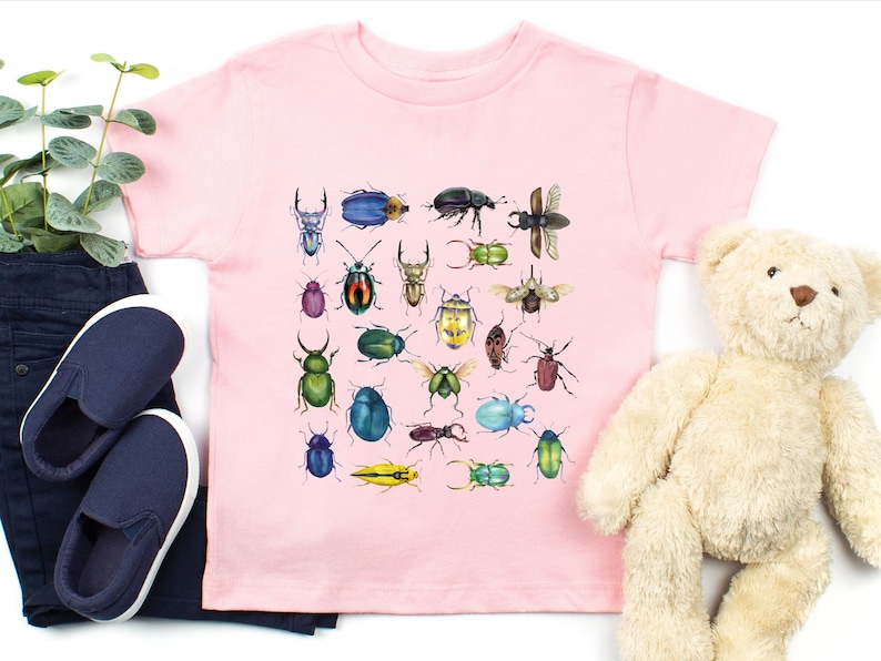 May include: A pink t-shirt featuring a colourful illustration of various beetles. The beetles are depicted in a range of colours, including green, blue, red, yellow, and black.