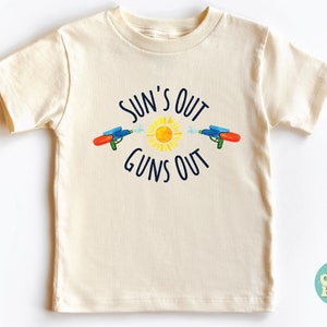 May include: Cream-colored t-shirt with the text "SUN'S OUT GUNS OUT" in navy blue. The design includes a yellow sun and two water guns in blue, red, and green. A fun summer-themed graphic tee for kids.