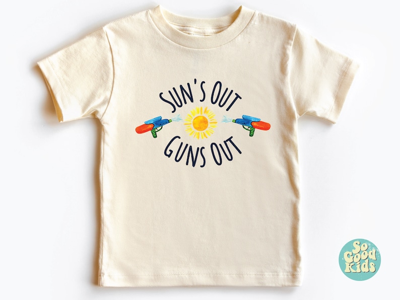 May include: Cream-colored t-shirt with the text "SUN'S OUT GUNS OUT" in navy blue. The design includes a yellow sun and two water guns in blue, red, and green. A fun summer-themed graphic tee for kids.